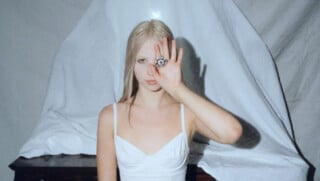 Das Foto zeigt ein Porträt von Stella Rose, die mit einer Hand ein Amulett vor ihr linkes Auge hält.