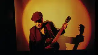 Das Foto zeigt den Musiker Matt Johnson von der Band The The, während er eine akustische Gitarre spielt.